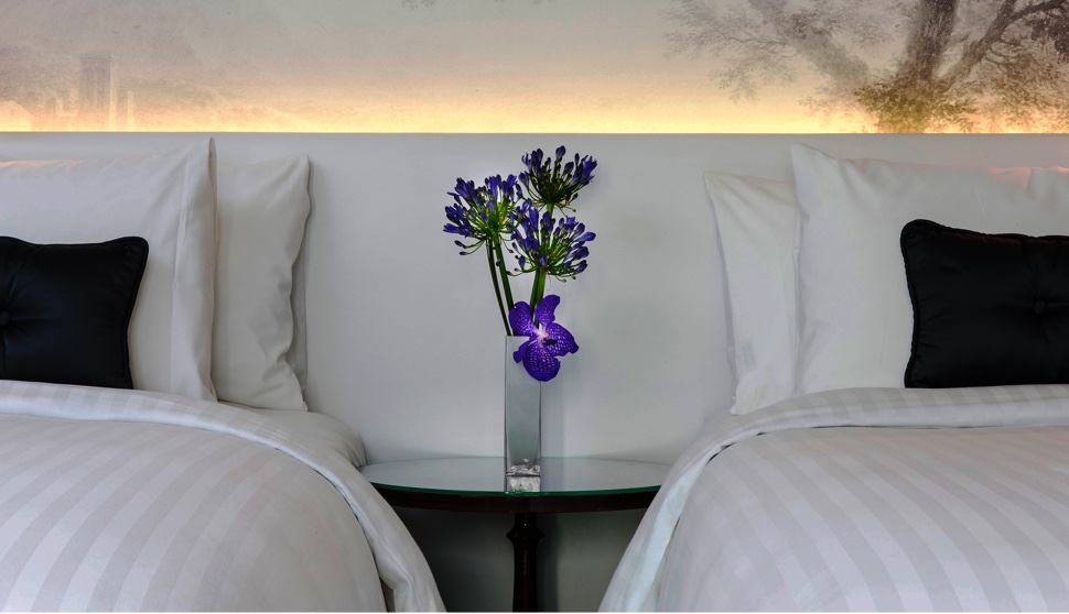 Two precisely made beds with a delicate floral arrangement on a table in between them.